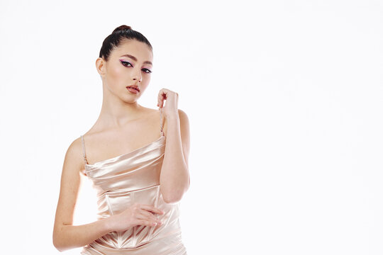 Elegant young woman in a silky beige dress poses with a thoughtful expression and arm bent. Soft makeup highlights her eyes and lips. Studio shot on a white background. Beauty fashion concept.