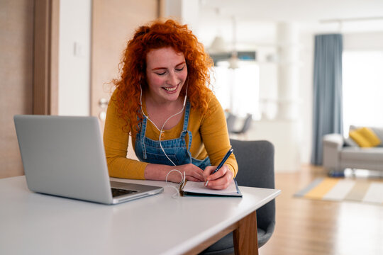 Young Red Haired Woman Smiling Indoors Wearing Yellow Sweater Using Laptop and Writing