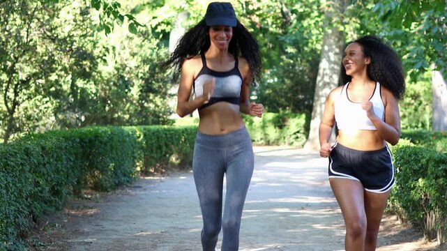 Multicultural Latin sportswomen jogging together along verdant park path, sharing fitness moment with infectious smiling and energetic stride