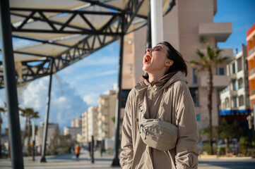 Fototapeta premium Young woman with an amazed open-mouth expression standing on the seaside promenade in Durrës during winter. Emotional lifestyle portrait. Surprise, joy, and spontaneous reaction 