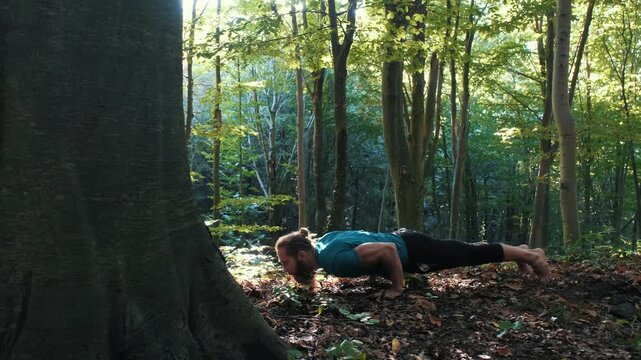 Bearded man practicing yoga in the forest, transitioning from plank pose to upward-facing dog (Urdhva Mukha Svanasana) during a morning flow.