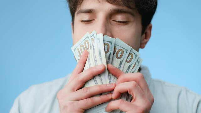 A person stands with closed eyes and smiles, holding multiple banknotes close to their face against a solid blue backdrop. Bright light highlights the scene.