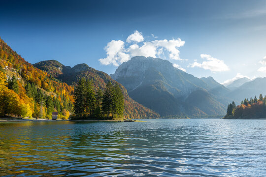 View of the tranquil lake mirroring the towering mountains under a bright sky, the trees displaying vibrant autumnal hues, Lago del Predil, Friuli-Venezia Giulia, Italy.
