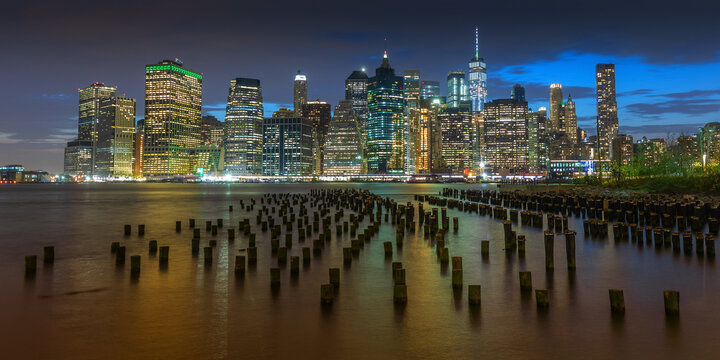 View of luminous skyscrapers pierce the twilight sky, reflecting in the shimmering water dotted with weathered wooden posts, a mesmerizing urban dance, New York City, USA.