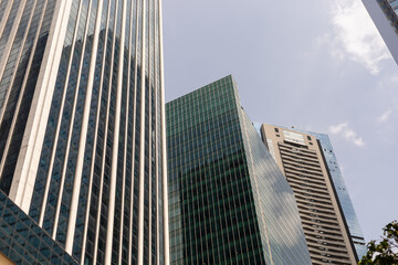 Obraz premium View of multiple tall buildings in a city. The structures show glass and steel designs. The sky is clear with a few clouds above. The scene captures urban life and architecture