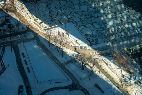View of stark white snow blankets the angular park pathways contrasting with the icy river reflecting the winter sun, New York, New York, United States.