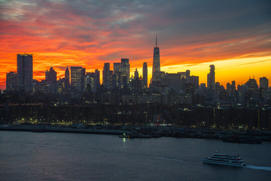 View of fiery sunset paints the sky above the silhouetted skyline and river, with a solitary boat gliding across the water, New York, New York, United States.