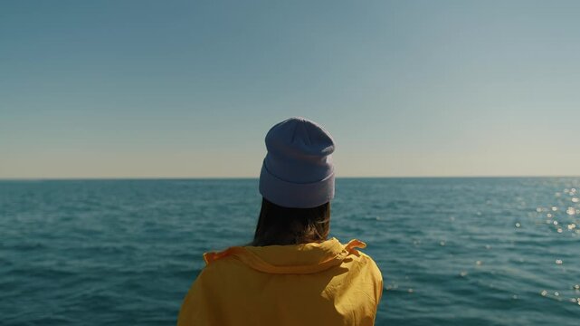 Kamchatka, Russia - August 26, 2025: Young girl in yellow jacket and lilac hat fishes with fishing rod from deck of yacht in open sea. Back view. Slow motion. Concept of silence and pacification