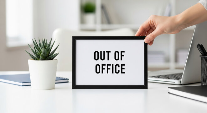 Close-up of hand placing a black frame with the text "Out of Office" on a white desk, signifying absence, break, or unavailable status