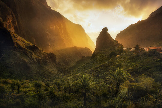 View of the village nestled within deep valleys with lush vegetation, dramatically lit by the sun, creating a serene yet vibrant scene, Masca, Canary Islands, Spain.