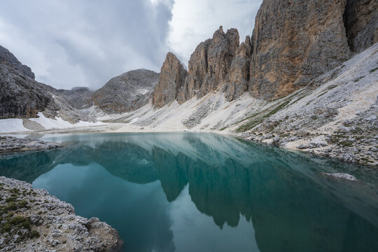 View of turquoise lake mirroring the jagged peaks and rocky slopes under a cloudy sky, a serene alpine scene in Valon de Antermoa, Campitello di Fassa, Trentino-Alto Adige, Italy.