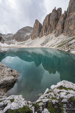 View of the tranquil turquoise lake mirroring jagged peaks under a soft, overcast sky, framed by moss-covered rocks in Valon de Antermoa, Campitello di Fassa, Trentino-Alto Adige, Italy.