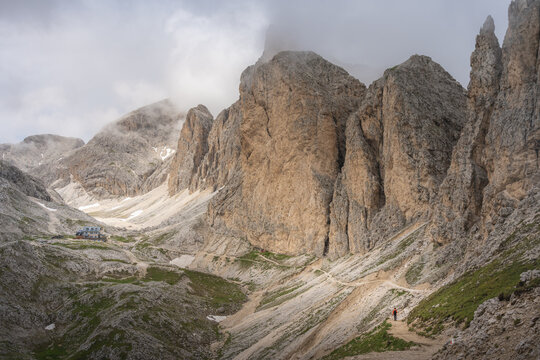 View of jagged Dolomite mountains reaching into a misty sky, with a small refuge nestled among the rocky slopes, inviting exploration, Campitello di Fassa, Trentino-Alto Adige, Italy.