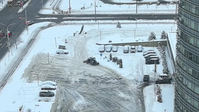 A snowplow works to clear a parking lot covered in snow. Cars are parked while snow piles up around them. The location is near a busy road in a city.