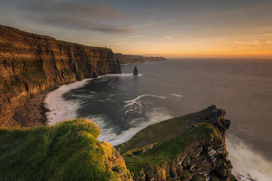 View of dramatic cliffs plunging into the churning Atlantic, where golden light kisses the rugged coastline, painting the scene in warm hues, Cliffs of Moher, County Clare, Ireland.