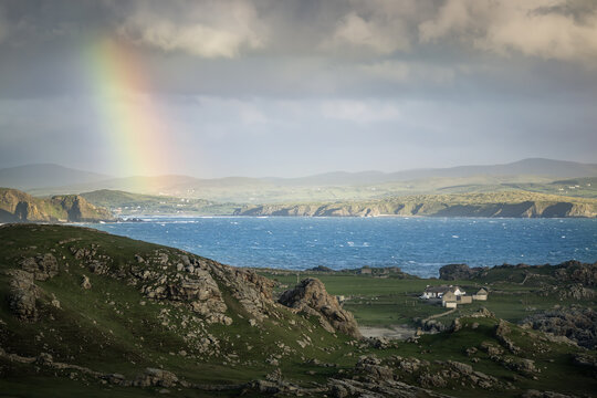 View of a vibrant rainbow arcs over the rugged coastline, with emerald hills meeting the turbulent blue sea, while small white buildings dot the verdant landscape, Malin Head, Ireland.