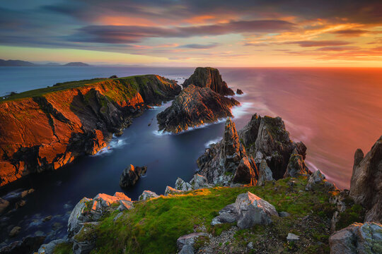 View of jagged cliffs meet the turbulent sea under a fiery sunset, painting the sky with hues of orange and purple, Malin Head, County Donegal, Ireland.