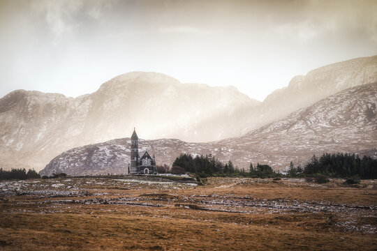 View of a quaint church nestled against the backdrop of majestic, snow-dusted mountains under a soft, diffused light in Dunlewey, County Donegal, Ireland.