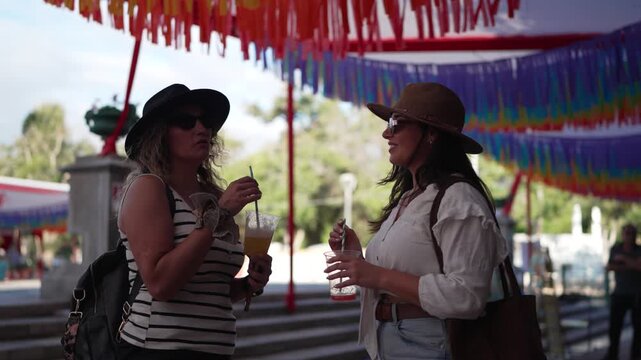 Two female friends enjoying drinks and conversation at a sunny outdoor city festival in La Serena 