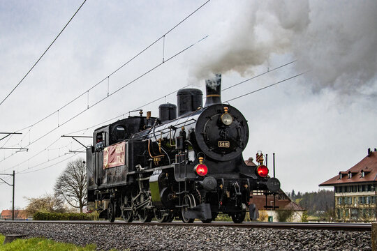View of a vintage black steam train billowing white smoke against a muted sky, chugging along the tracks near quaint buildings, Lyss, Canton of Bern, Switzerland.