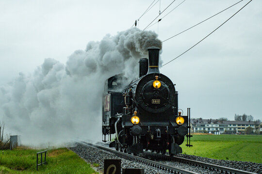 View of a vintage steam engine billowing smoke, its golden lights gleaming against the green fields and overcast sky, Lyss, Canton of Bern, Switzerland.
