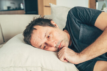 Depressed mature man lying on couch with sad expression, struggling with emotional pain, loneliness, or mental health issues in a quiet home interior.