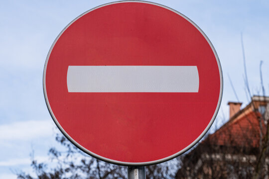 a circular red traffic sign with a horizontal white bar&mdash;universally recognized as "No Entry"&mdash;set against a blue sky with blurred trees and a building roof.