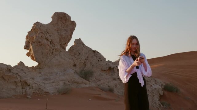 Young caucasian woman in white shirt standing on sand dunes in desert while using smartphone at sunset, representing travel, digital communication and modern lifestyle.