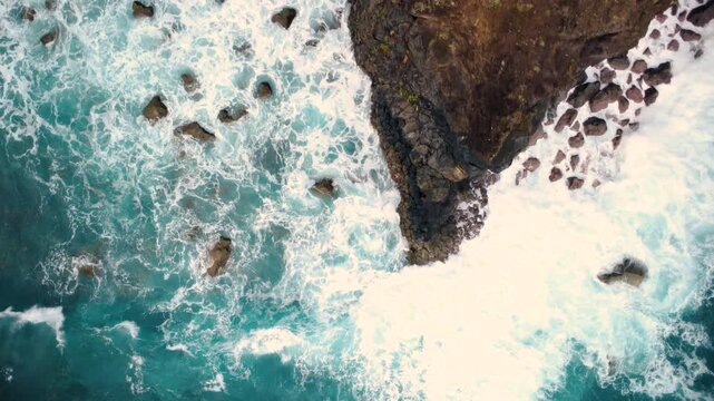 Aerial top down view of a sharp rock cliff edge, drone flying forward from the ocean towards the coast revealing crashing waves and white foam at Miradouro do Guindaste in Madeira island.