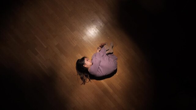 Little girl lying curled up on a wooden floor, spotlight highlighting her presence in a dark room, capturing feelings of vulnerability, anxiety, and solitude
