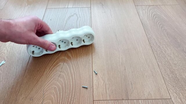 A hand uses a screwdriver to close a white three - socket power strip on a wooden floor, revealing screws and components, demonstrating household electrical repair and maintenance work