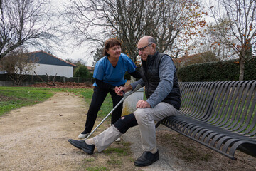 Caregiver helps elderly man sit on bench in a park setting