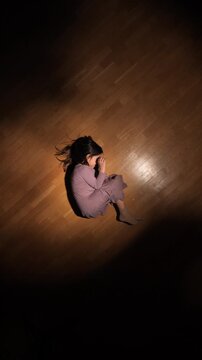 Vertical video young girl sitting on a wooden floor in a spotlight, hiding her face while experiencing overwhelming sadness, feeling lonely and vulnerable in the darkness