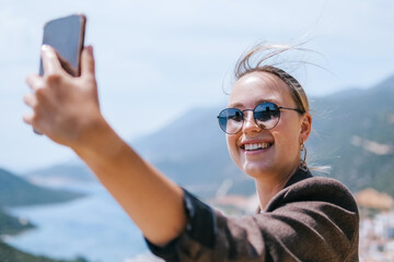 Travel blogger filming live stream on smartphone against mountain coastline, Gen Z female tourist on viewpoint, panoramic sea and hills, social media content creator, sunny outdoor vacation vibe. © shintartanya