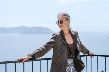 Travel blogger filming live stream on smartphone against mountain coastline, Gen Z female tourist on viewpoint, panoramic sea and hills, social media content creator, sunny outdoor vacation vibe. © shintartanya