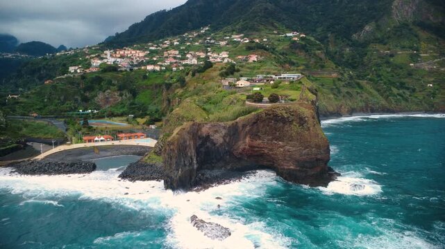 Drone flying backwards with a turn revealing Miradouro do Guindaste and Faial village, aerial view of coastal cliffs, Atlantic ocean waves and green mountains under cloudy sky in Madeira island.