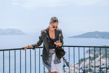 Travel blogger filming live stream on smartphone against mountain coastline, Gen Z female tourist on viewpoint, panoramic sea and hills, social media content creator, sunny outdoor vacation vibe. © shintartanya
