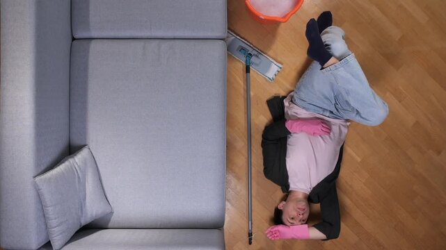 Exhausted person wearing protective gloves lying on a hardwood floor, taking a break from cleaning, with a mop and bucket nearby, feeling tired and overwhelmed by household duties