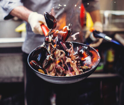 Close up of chef tossing mussels with vegetables in frying pan over open flame. Dynamic restaurant cooking process, seafood cuisine action shot.