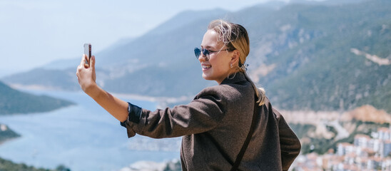 Travel blogger filming live stream on smartphone against mountain coastline, Gen Z female tourist on viewpoint, panoramic sea and hills, social media content creator, sunny outdoor vacation vibe. © shintartanya