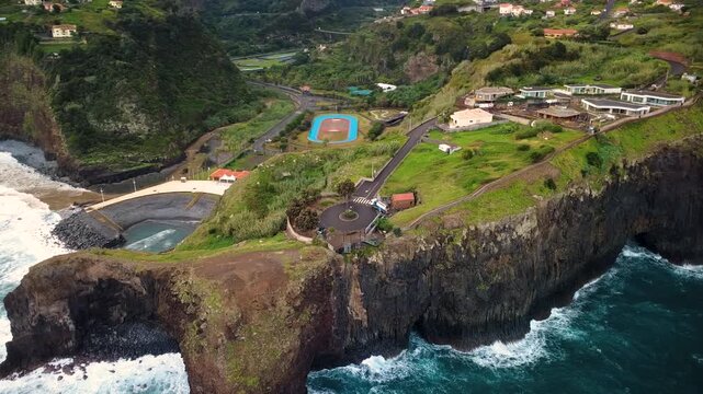 Drone orbit shot of Miradouro do Guindaste in Madeira, aerial view of Faial coastline with high cliffs, sports ground, village houses and Atlantic ocean waves under cloudy sky on northern coast.