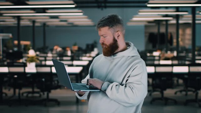 The man stands in an office environment using a laptop. The space features multiple desks with bright overhead lights and no people around.