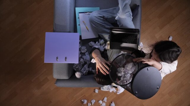 Child in home office scattering crumpled paper from a trash can while father struggles to work on his laptop, captured from overhead chaos, distraction, work life tension