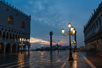 Fototapeta premium Empty Piazza San Marco in Venice at dawn, with glowing street lamps reflected on wet stone pavement after rain. Historic buildings and columns create a dramatic and cinematic atmosphere before sunrise