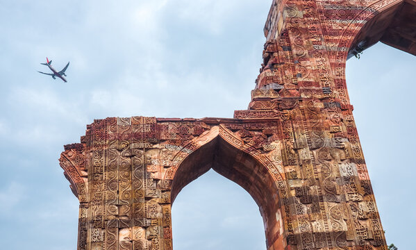Modern commercial airplane flying over ancient ruins of Qutub Minar complex in Delhi India with intricate sandstone carvings and historic Islamic architecture under overcast sky during summer day