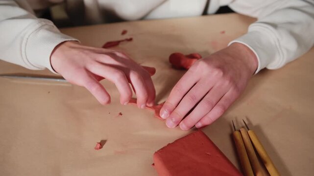 Detailed shot of hands meticulously shaping red modeling clay. The artist uses small tools to sculpt and craft the pliable material on a brown paper surface, showcasing creativity and skill.
