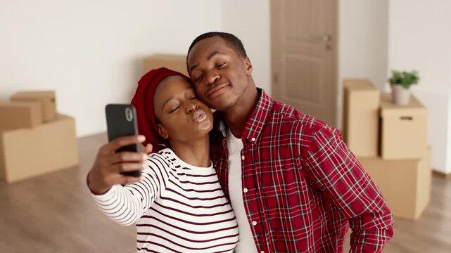 A woman with dark skin and a man with dark skin pose together while taking a selfie in their new home. They are surrounded by unpacked boxes, showing excitement during the moving process.