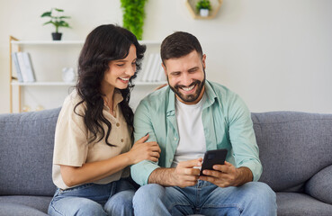 Obraz premium Happy couple using smartphone together at home. Smiling man and woman sitting on sofa in living room to look on screen, make online shopping order and payment for purchases, video call to friends