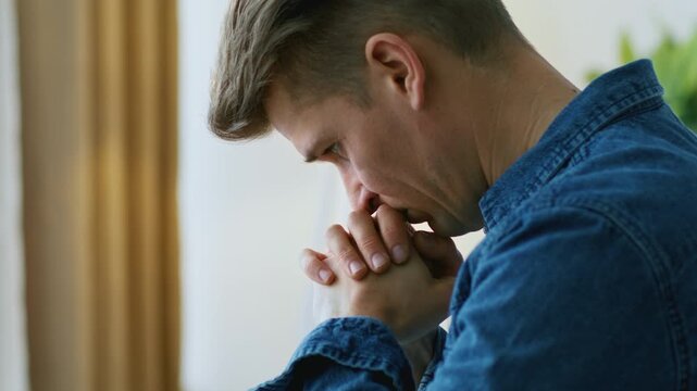 A man is sitting indoors, hands clasped together, appearing deep in thought. Natural light enters the room creating a bright atmosphere. He is focused and contemplative.