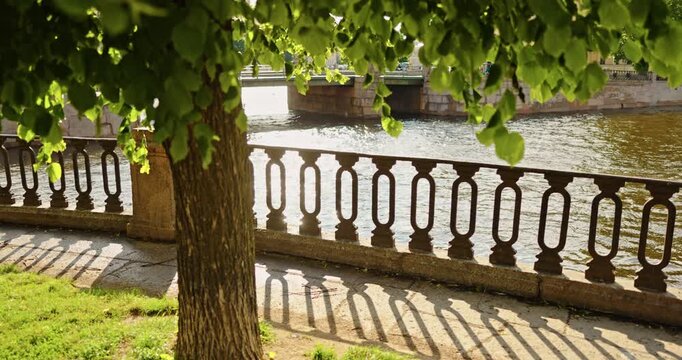 a promenade with wrought iron railings through green leaves swaying in the wind near St.Nicholas cathedral in summer day, the embankment of the Kryukov Canal and the Griboyedov Canal
